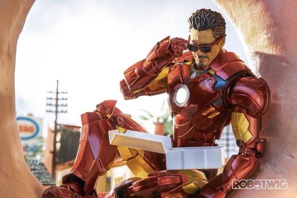 Tony Stark sits in a giant doughnut eating a box of doughnuts in his iron man suit without his helmet. He wears sunglasses and is pulling them down as someone below is talking to him