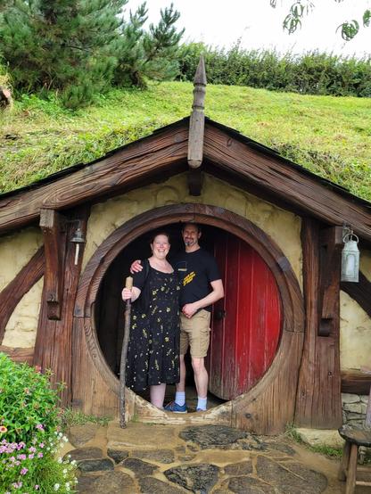 William and Heather at the entrance to a hobbit hole with a red door.
