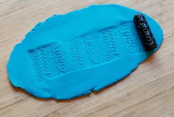 A black cylinder is rolled across a flattened piece of bright blue clay, leaving behind a repeating pattern of geometric shapes and symbols.
