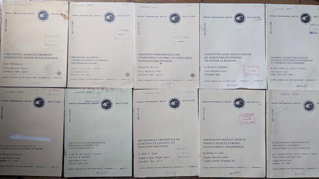 A photo of a bunch of NASA research documents, with titles like "STRUCTURAL MARGINS OBSERVED IMMEDIATELY PRIOR TO EXCEEDANCE", "PRELIMINARY ANALYSIS OF UNEXPECTED WOBBLE IN A RIGIDLY NON-WOBBLING SYSTEM", "OBSERVED PERFORMANCE OF COMPONENTS KNOWN TO HAVE BEEN INSTALLED BACKWARDS", "COGNITIVE LOAD IMPLICATIONS OF ACRONYMS REFERRING TO OTHER ACRONYMS", "THRMAL CHARACTERISTICS OF MATERIALS DESCRIBED AS 'PROBABLY INFLAMMABLE'", "ON THE REPEATED SURVIVAL OF SYSTEMS NEVER INTENDED TO WORK", "THE EFFECT OF CLIPBOARD ON PERCEIVED AUTHORITY IN TEST ENVIRONMENTS", "MECHANICAL PROPERTIES OF SUBSTANCES EXPOSED TO REPEATED MEETINGS", "ANOMALOUS RESULTS WHICH PERSIST DESPITE STRONG MANAGEMENT DISAPPROVAL", and "A PRELIMINARY ASSESSMENT OF THE MOON'S APPARENT DISINTEREST IN OUR ACTIVITIES"