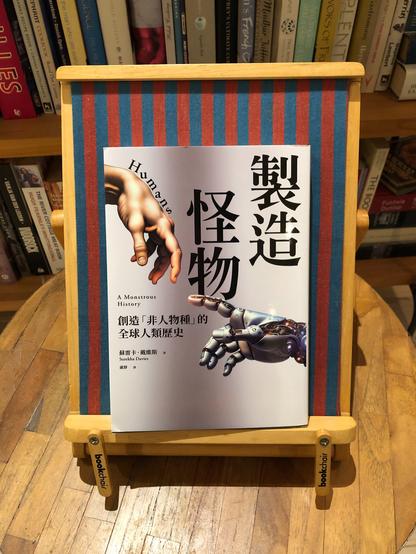 A pale grey softcover book on a red and blue striped book chair on a circular wooden coffee table, with bookcases in the background. The book cover: black text in complex Chinese characters of various font sizes. In smaller font in Latin characters are "A Monstrous History" and "Surekha Davies". At the top left and bottom right are two large, outstretched hands, human (top) and silvery-grey robot with some exposed machinery (bottom), reminiscent of the hands of God and Adam on Michelangelo's Sistine Chapel ceiling.