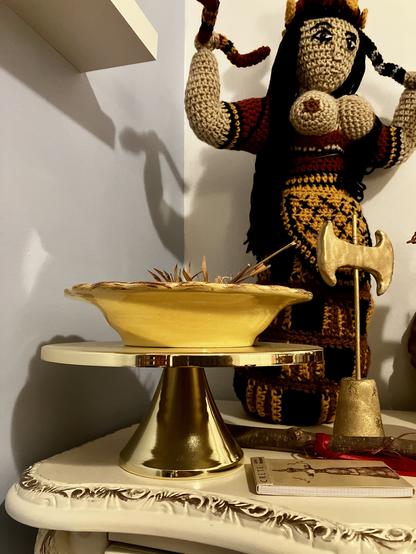 A view of one corner of one of my altars, in this case a Minoan one. In front is a shiny gold footed cake plate topped with a yellow ceramic bowl containing some small dried palm fronds. Behind this is a tall crocheted copy of the Minoan Snake Goddess figurine. To the right is a small gold labrys in a stand, the handle of a sistrum with red ribbon wrapped around it, and a small notepad with a photo of the Snake Goddess figurine on the cover.