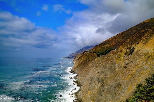 View of the California coast, looking north through tree branches from Ragged point. There is surf breaking on the shore. The sky is cloudy.