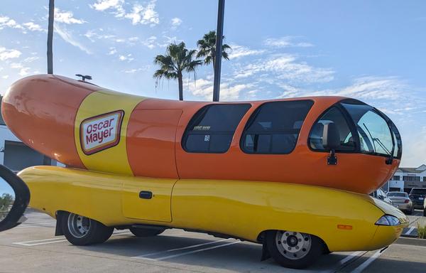 A car shaped like an upward-curving hot dog on a wavy base so it looks like a giant hot dog has been smooshed onto a long 1950s convertible. The base is yellow, the hot dog is bright orange, and there are curved windows set into the front so a driver can see where they're going. The Oscar Mayer logo is painted on the side, looking like the strip of labeling in their typical hot dog package. It's all bright and shiny, clearly well-maintained and not just a relic of a bygone era.