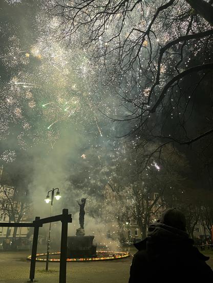 Eine Statue steht in einem Park, umgeben von bunten Laternen, mit einer Person, die ein lebendiges Feuerwerk beobachtet, das den Nachthimmel erhellt. Rauch und Funken erfüllen die Luft und schaffen eine festliche Atmosphäre.