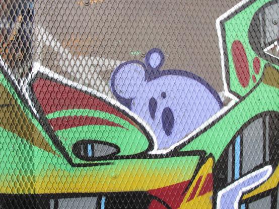 Bold graffiti with neon green, maroon, yellow, and lilac purpose in vaguely organic but geometric shapes over a plywood wall that is covered with a perforated metal sheet in a diamond pattern.