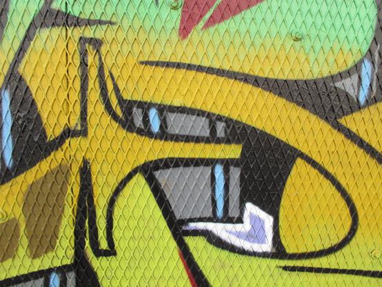 Bold graffiti with neon green, maroon, yellow, and lilac purpose in vaguely organic but geometric shapes over a plywood wall that is covered with a perforated metal sheet in a diamond pattern.