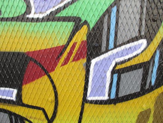 Bold graffiti with neon green, maroon, yellow, and lilac purpose in vaguely organic but geometric shapes over a plywood wall that is covered with a perforated metal sheet in a diamond pattern.