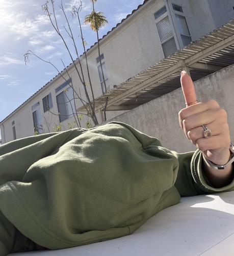 Woman with her head buried in her hood, giving a halfhearted thumbs up after she drug herself outside against her own will.