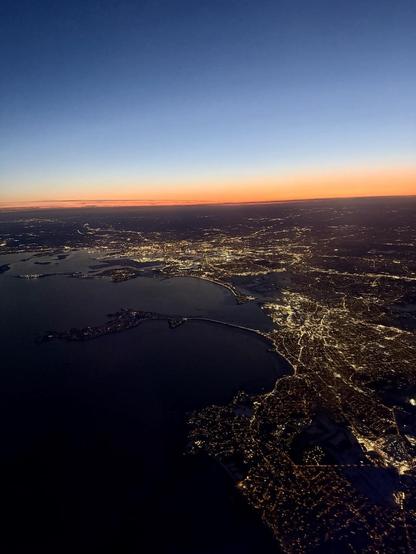 High altitude view of a darkened harbor. The ground is lit up with a network of interconnected lights.  The sky is a dark orange near the horizon with a narrow band of golden yellow above that and darkening blue above that.