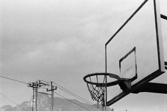 NEOPAN SS 100 (FF)

English Alt Text:
A black-and-white photo of an outdoor basketball hoop mounted on a rectangular backboard with a marked square target. The hoop has a net and stands against a cloudy sky. In the background, utility poles and power lines stretch across the scene, with faint hills or mountains visible in the distance. The image evokes a quiet, nostalgic mood, emphasizing simplicity and open-air recreation.
中文替代文字：
一张黑白照片，展示一个户外篮球框，安装在一个带有方形标记的矩形篮板上。篮筐挂着网，背景是多云的天空。远处有电线杆和电线横穿画面，隐约可见山丘或山脉的轮廓。画面营造出宁静怀旧的氛围，突显简约和户外运动的意境。