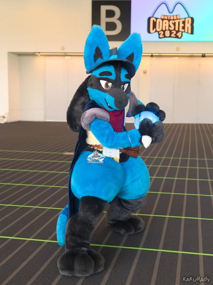 A person dressed in a Lucario costume — a blue canine with tan body and black feet, and black hair-like appendages behind the head — wearing a red scarf, a blue cape and a blue pointed hat, standing on a brown carpet with stripes, holding up a glowing ball to the side and towards the back of their body, as if about to throw it. In the backround are several gates that are closed, above which there is a video signage that displays "Anthro Coaster 2024".