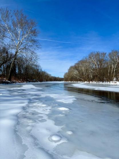 A broad creek flows between wooded areas under a clear blue sky. Thick ice and snow cover most of its surface, while the center remains capped with thinner, glassy ice that lets the dark water show through.
