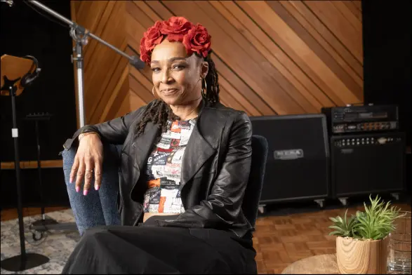 Siedah Garrett seated in a recording studio with one arm perched over the back of a blue chair. She is a wearing a red flowered hair decoration, a white shirt with graphic patterns, a black leather jacket, and black pants or long skirt. She is smiling slightly. Photo Credit: Sekou Luke