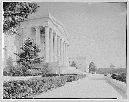 The image shows a black and white photograph of the National Gallery, captured from its front view with the United States Capitol building visible in the background. The gallery's neoclassical architecture is prominently displayed, featuring tall columns and large windows framed by classical moldings. In the foreground, there are neatly trimmed bushes on either side of a paved walkway leading towards the entrance of the National Gallery.
The trees surrounding the area appear to be young due to their slender trunks, suggesting that this photograph might have been taken at an early stage in its establishment or after recent landscaping efforts. The overall setting appears serene and well-maintained with clear skies above, indicating a pleasant day for visitors to enjoy the art institution's offerings.
This image is credited to Theodore Horydczak (1890-1971) as indicated by his name in the description provided. Additionally, it mentions that this particular negative measures approximately 691mm x 692mm and has a resolution of safety glass with dimensions around 8 inches on each side. The source information for this photograph is available at https://images.loener.nl/horydczak/full/691e/691ee665450e57a2674dc6ed.jpg, suggesting that it may be part of a larger collection or archive pertaining to Horydczak's work.