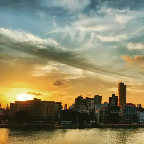 Céu durante pôr-do-sol no Recife. em primeiro plano, o Rio Capibaribe, com os prédios do bairro de São José ao fundo. Atrás, o céu com o sol na altura do horizonte, mais amarelado e tornando-se azul mais acima.