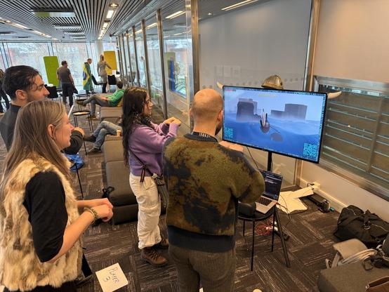 Four people stand in front of a TV screen which has a boat in an ocean