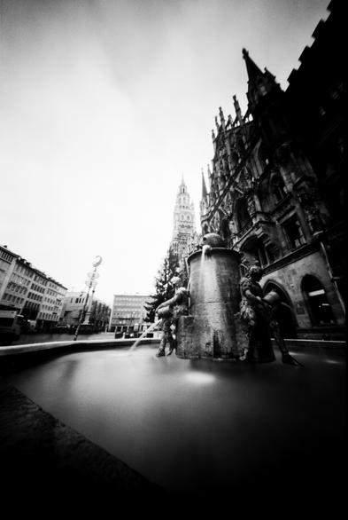 The black and white image shows the Fischbrunnen (Fish Fountain) in the Marienplatz square of Munich, Germany, with the distinctive Gothic Revival architecture of the New Town Hall (Neues Rathaus) towering in the background. The image was taken using a pinhole camera, giving it a distinctive dreamy, soft focus and infinite depth of field.