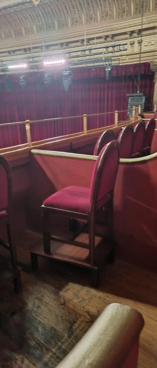 Primer plano de los asientos en el palco superior del teatro. Se trata de taburetes altos de madera con respaldo y asiento tapizados en terciopelo rojo. Estos taburetes están colocados sobre una estructura o tarima de madera adicional para ganar altura respecto a la barandilla del palco. Al fondo se ve la barandilla de seguridad y el telón rojo del escenario, mostrando la perspectiva lateral y elevada del espectador.
