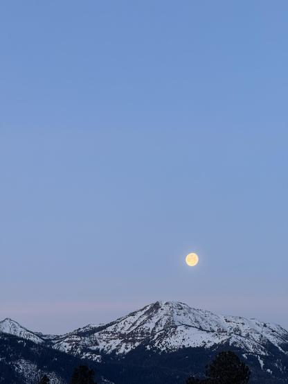 The bright, nearly full moon hangs in the clear, pale blue morning sky over snow-capped mountains as it heads down toward the western horizon