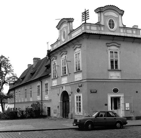 Architettura: L'edificio principale è una costruzione d'epoca a due piani (più il tetto), situata all'angolo di una strada. Ha uno stile barocco o rococò, con due frontoni decorativi sul tetto che ospitano finestre ovali. Sulla facciata si legge chiaramente il cartello stradale "LORETÁNSKÁ".

Dettagli stradali: Al piano terra c'è un portone ad arco scuro e un ingresso laterale con la scritta "GALERIE".

Veicoli: Davanti all'edificio è parcheggiata una berlina squadrata, tipica degli anni '80 o inizio anni '90 (probabilmente una Škoda), che dà un contesto temporale alla foto. La strada è fatta di sanpietrini.