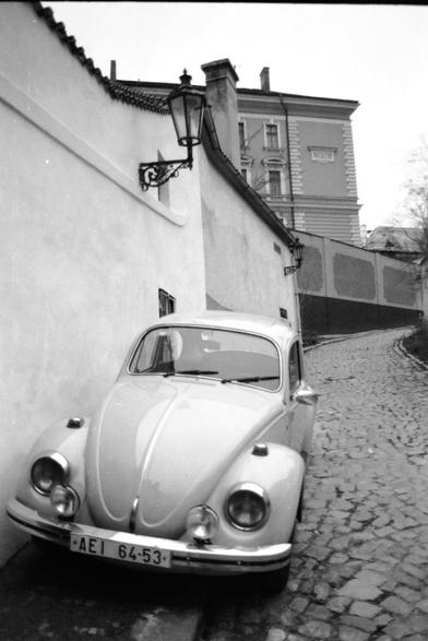 Il Maggiolino nel Vicolo Questa foto è un primo piano di un'auto d'epoca parcheggiata in una strada molto stretta.

Soggetto: Un Volkswagen Maggiolino (Beetle) classico di colore chiaro domina la parte bassa dell'immagine. Si vedono bene i fari rotondi, il paraurti cromato e la targa "AEI 64-53".

Ambiente: L'auto è parcheggiata accanto a un alto muro bianco sulla sinistra.

Dettagli architettonici: Sul muro è fissato un antico lampione in ferro battuto dal design elegante. La strada è in forte pendenza e lastricata. Sullo sfondo si vede un grande edificio a più piani con finestre classiche, che chiude visivamente la scena.
