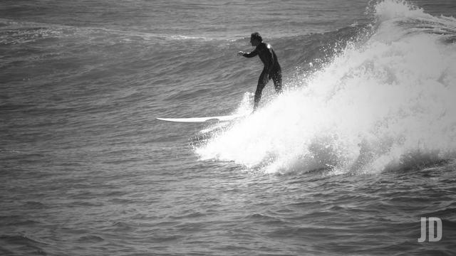 La imagen en blanco y negro muestra a un surfista montando una ola. El surfista, con traje de neopreno, se encuentra de pie sobre la tabla mientras la ola rompe detrás de él, generando espuma y salpicaduras en el agua. El mar ocupa la mayor parte de la escena, transmitiendo movimiento y dinamismo.