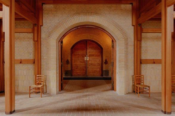 Interior of a spacious hall featuring wooden beams, arched doorways, and two simple wooden chairs against a light brick wall.