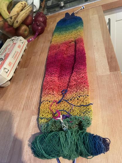 An in-progress knitting project on a wooden kitchen surface. From the far end a mirror rainbow gradient starts at purple (which is ribbing) then goes into blue as the lacework starts, through green and yellow where the piece splits from in the round to flat. Orange, pink, pink, orange, yellow, green.