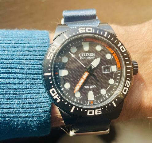 A black dial dive watch from citizen, with some orange highlights on the minute hand and the arc from 60 to 15min. Mounted on a deep blue nato strap. Worn on a Caucasian man’s wrist, wearing a teal sweater.