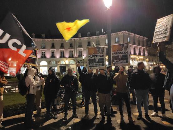 Photo d'une partie du rassemblement pour le Rojava, un drapeau UCL à gauche devant une pancarte UCL Kurdistan, en haut un drapeau des YPG et en dessous deux pancartes UCL Kurdistan et à droite la 4è pancarte Kurdistan