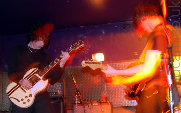 Two men on a stage playing electric guitars as part of a rock band, the man on the left more or less facing the camera, the other facing off to the left.
The man on the left is wearing a long-sleeved top, with a chain dangling from round his neck, and dark trousers, playing a cream coloured electric guitar.
The man on the right is wearing a black t-shirt and black jeans, and playing a black electric guitar.
In the background, industrial metal floor plates are mounted on the purple walls, behind stacks of amplifiers, with a purple ceiling.
A flash has frozen both people in place, while a long exposure has caught movement, producing yellow and red secondary images.