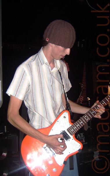 A man wearing a brown-striped white shirt and faded blue jeans, and a woolly hat on his head, playing an orange electric guitar.
He is looking down towards his instrument as he plays.
In the background, black drapes mostly cover brown brick walls, with amplifiers visible behind him on the left, and someone playing a bass guitar on the right.