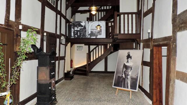 The image depicts the interior of a traditional half-timbered building, likely historical in nature. The walls are characterized by exposed wooden beams with white plaster in between, giving the space a rustic and vintage charm. The floor is covered with small, patterned tiles.
In the center of the room, a wooden staircase leads to an upper level, which features a railing and additional display space. On the upper level, there are black-and-white photographs mounted on the wall, depicting historical scenes or figures.
On the lower level, to the right, there is a large framed portrait of a woman dressed in historical attire, possibly from the late 19th or early 20th century. The portrait is propped up on an easel.
To the left, there is an old-fashioned, ornate cast-iron stove with decorative elements, and a green plant placed beside it, adding a touch of natural color to the room. The overall atmosphere is one of historical preservation, evoking a sense of nostalgia and curiosity about the past.