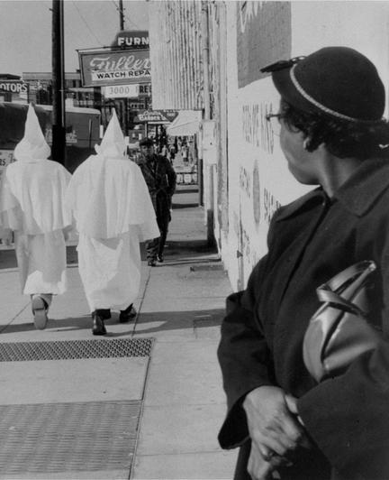 Two robed and hooded Klu Klux Klan members walking down a Montgomery AL sidewalk the night before a cross-burning, after a permit to parade in the street was denied. In the foreground, a woman of color watches.