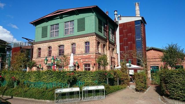 The image shows a charming, historic industrial building that has been repurposed. The building features a mix of architectural styles, with a lower section made of red brick and an upper section painted in a light green color with white window frames. The roof is slightly sloped and has a traditional design.
Attached to the building is a tall, narrow tower-like structure with a rusty, reddish-brown exterior and vertical windows, possibly an old chimney or industrial stack.
In front of the building, there is a well-maintained garden area with green bushes and plants, adding a touch of nature to the scene. There are also outdoor seating areas with umbrellas, suggesting a café or restaurant space. Bike racks are visible in the foreground, indicating that the area is accessible and frequented by visitors.
The sky is clear and blue, suggesting a bright, sunny day, which enhances the overall inviting and pleasant atmosphere of the location.