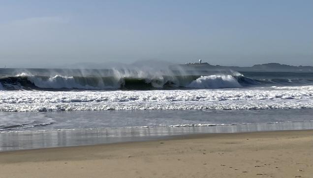 A long line of waves breaking near a sandy beach, and throwing up a lot of spray. The sky is a cloudless blue.