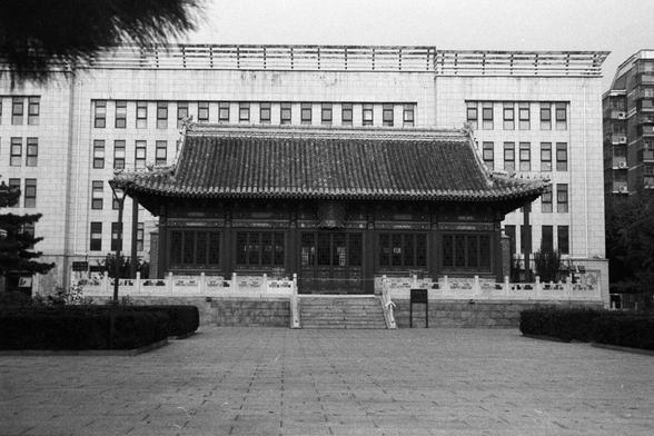 NEOPAN SS 100 (FF)

Image 2 – English Alt Text
A traditional Chinese building with ornate roof tiles, upturned eaves, and carved wooden details stands on a raised stone platform with steps leading up. In the background, a modern multi‑story building with a grid of identical windows rises behind it, creating a contrast between old and new architecture. The foreground shows a paved courtyard with trimmed hedges and a simple lamppost.
Image 2 – 中文替代文字
一座传统中式建筑立于石台上，屋顶有飞檐和精致瓦片，木结构雕刻细节清晰。台阶通向入口。背景是一栋带规则窗格的现代高层建筑，形成古今建筑的对比。前景是铺装庭院，有修剪整齐的绿篱和一根简洁的路灯杆。
