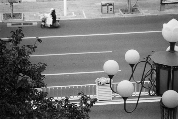 NEOPAN SS 100 (FF)

Image 1 – English Alt Text
A black‑and‑white overhead view of a wide, mostly empty street. A single person rides a scooter in the center lane, carrying several large bags or bundles stacked behind them. The road markings are clean and geometric. On the sidewalk to the left, there are two trash bins, a bicycle‑lane symbol, a tall pole, and several young trees planted in square grates. In the foreground, an ornate streetlamp with multiple spherical bulbs stands beside a planter filled with flowers and a metal barrier. The scene feels quiet and spacious.
Image 1 – 中文替代文字
黑白俯视街景。一名骑电动车的人行驶在宽阔、几乎空无一人的车道中央，车后堆放着几个大包裹。道路标线清晰。左侧人行道上有两个垃圾桶、自行车道标志、一根立杆和几棵种在方形树池里的小树。前景是一盏多球形灯头的装饰路灯，旁边有花箱和金属护栏。整体画面安静而开阔。