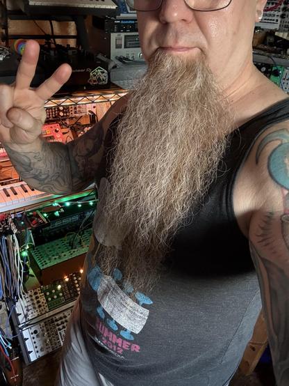 A person with a long beard and tattoos is taking a selfie while making a peace sign. They are wearing a tank top with a graphic design that looks like the Woodstock festival logo. In the background, various music equipment and synthesizers are visible, illuminated by colorful lights.