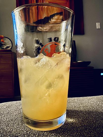 In a pint glass is a yellow-orange liquid and some ice cubes.