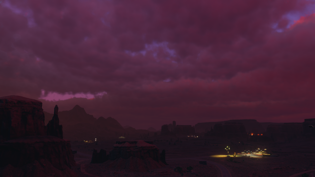 Night shot of a rural desert road with a gas station illuminated by warm streetlights. Numerous buttes and mesas dot the arid landscape. The clouded sky hosts shades of purple, pink, apricot, and orange.