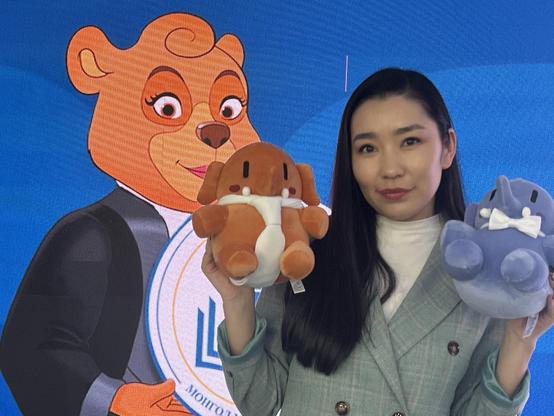 Frau in grünem Blazer mit unseren beiden Plüschelefanten in beiden Händen, im Hintergrund auf einer digitalen Anzeige ein großer Bär im Comic-Stil, das Logo der Justizakademie der Mongolei.