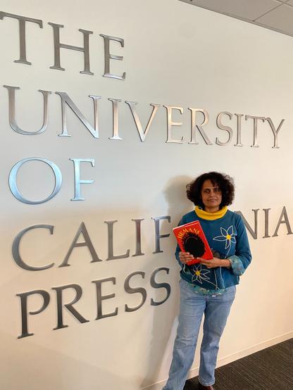 Me in a blue and yellow sweater and blue jeans, holding a copy of HUMANS, standing against a white wall with silvery lettering spelling out "University of California Press", looking dazed. - At the UC Press head office.