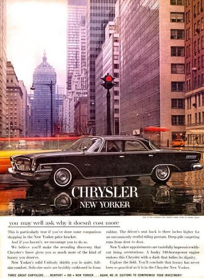 The image is a full-page vintage magazine advertisement for the Chrysler New Yorker automobile. A long, black four-door sedan is shown in profile, centered in the image, stopped or moving slowly through a wide city street. The pavement is wet, creating a reflective surface beneath the car.

The setting is a dense urban downtown, identified as New York City. Tall office buildings line both sides of the street, forming a canyon of glass and stone. In the distance, a prominent skyscraper with a distinctive crown rises above the surrounding buildings. A traffic signal above the car shows a red light. Blurred streaks of red and yellow lights suggest passing traffic and motion around the stationary vehicle.

The car itself is large and low, with chrome trim along the sides, prominent wheel covers, and whitewall tires. Its long body and formal roofline emphasize luxury and size.

Centered beneath the image, large capital letters read “CHRYSLER NEW YORKER.” Below this headline is a block of smaller text beginning with the line “you may well ask why it doesn’t cost more,” followed by several paragraphs describing the car’s comfort, interior features, ride quality, and value. The text emphasizes luxury, quietness, power, and practicality.

At the very bottom of the advertisement is a narrow line of additional promotional text referencing other Chrysler models.