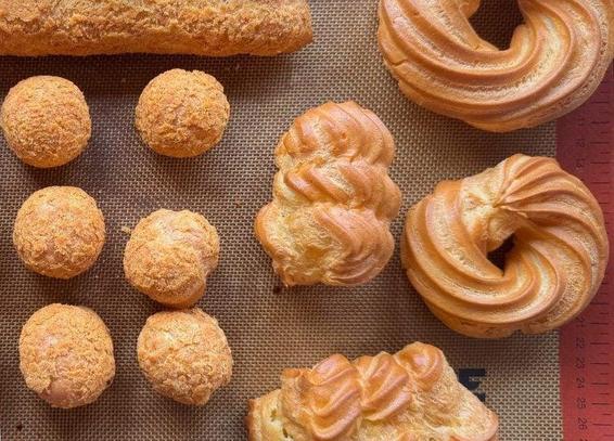 An overhead shot shows various golden-brown choux pastries, including round cream puffs and ring-shaped Paris-Brest, arranged on a textured brown baking mat.