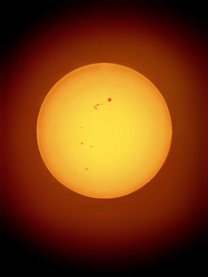 Photo of the sun as seen through a telescope with a solar filter. It appears as a bright yellow ball surrounded by a soft red-orange glow that fades to black at the edges of the image. A group of prominent sunspots can be seen at the center of the sun’s northern hemisphere, with a scattering of other sunspots below.

Both photos in this post were shot on an iPhone 15 attached to a Celestron 130mm reflector telescope with a Celestron EclipSmart orange-tint solar filter. Edited in Snapseed to bring out more contrast and structure in the sunspots.