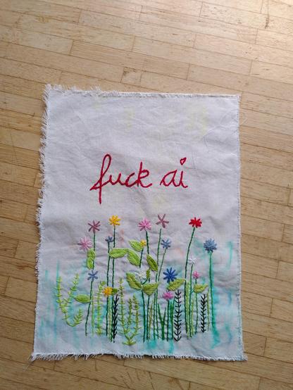 The same embroidery piece as in previous posts of this thread, now finished.

On a piece of off-white muslin cloth, it says "fuck ai" in the top half of the fabric, stitched in cursive with red yarn in chain stitch.
Below, a flower meadow with multi-colored flowers, some green leaves, some branches with tiny leaves, flower stems and grass. Washed-out greenery (done with textile markers) is visible in the background.