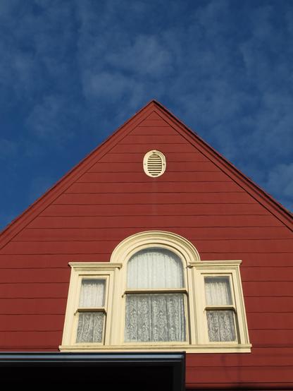 Tip top of a redish house against a dark blue sky. It has very similar windows as the first photo, with the center having a round top, but there are no leaded patterns on the side. There is minimal trim along the roofline, but there is an oval vent for the roof at the peak.