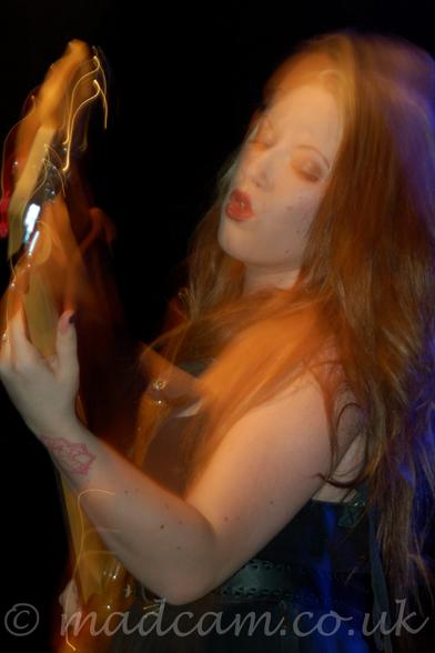 A long exposure close-up photo from the side of a woman with long blonde hair cascading over her shoulders, wearing a black dress that leaves her arms and shoulders bare, playing  a brown bass guitar.
She is leaning slightly backwards, and is looking at her left hand.
There is a red tattoo on the back of her left wrist.
A flash has frozen her in place, with streaks of lights showing how she has moved afterwards.
The background is entirely black.